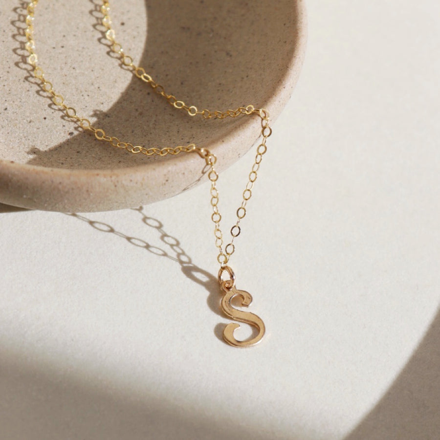 14k gold fill initial Script Necklace laid on a gray plate in the sunlight. 