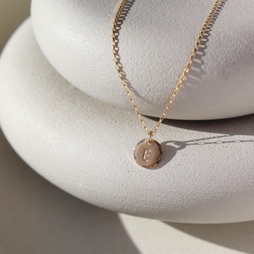 Small Monogram Coin Necklace laid on two white stone plates in the sunlight. This necklace features the coin chain followed by the mini coin tag that is hammered on the upside and is marked with the initial E.