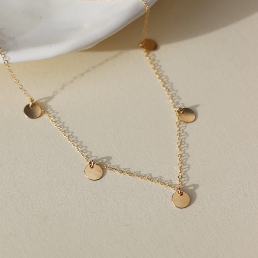 14k gold fill Suncatcher Necklace laid on a tan paper in the sunlight. This Necklace features the simple chain with 5 circle disc. This necklace is perfect for layering.