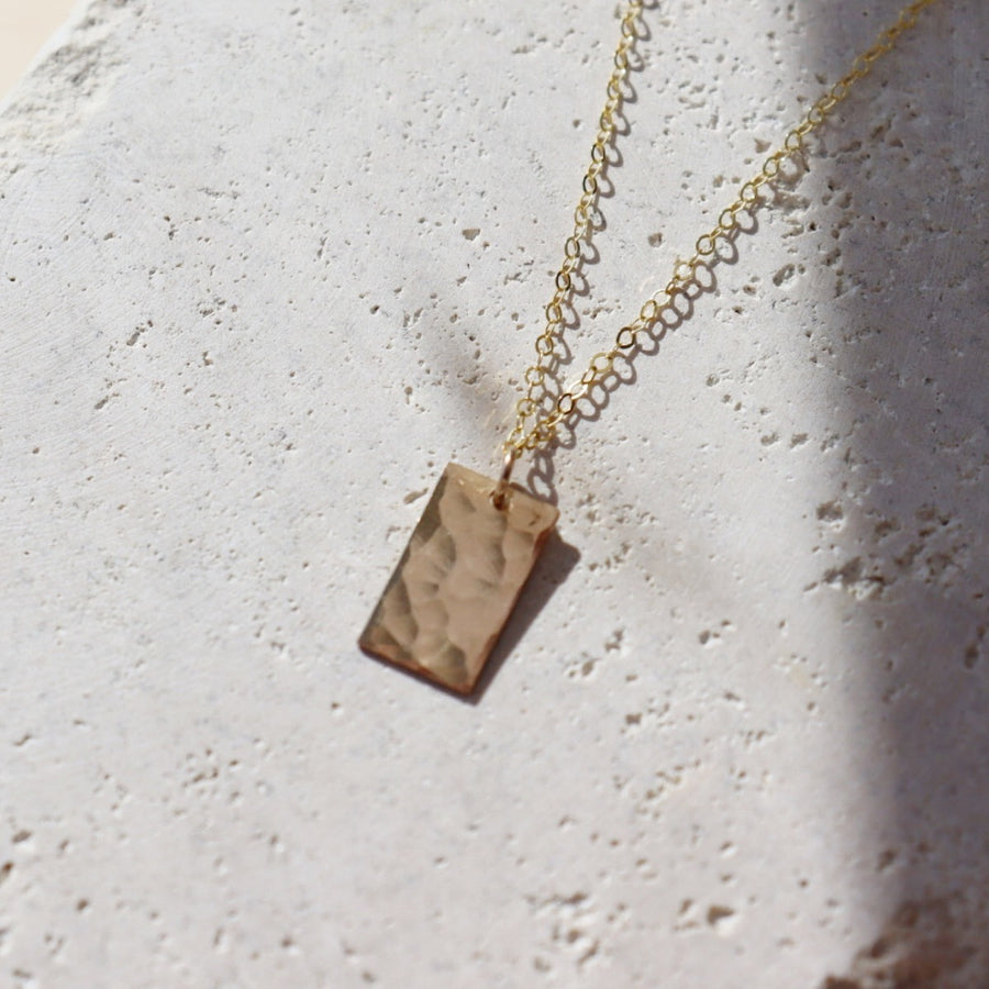 Rectangle 14k gold filled charm suspended from a delicate cable chain with clasp closure. Charm is hammered to a pretty shine and displayed on a white plate. Handmade by Token Jewelry in Eau Claire, WI Box Charm Necklace - Token Jewelry Edit alt text