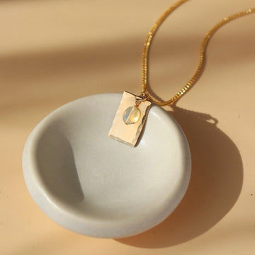 14k Everly Charm Necklace laid on a white ring dish in the sun. This necklace feature a simple chain a box charm with hammering on the outside is connected along with a opal stone. - Token Jewelry 